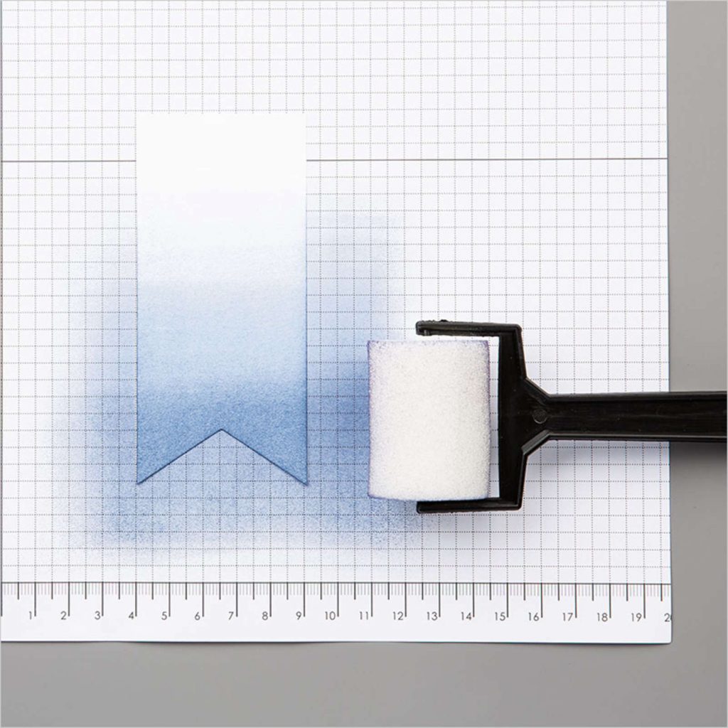 White grid paper with a white sponge cylinder attached to a black handle used to roll a blue ink.  Image shows how to use sponge brayering for cardmaking