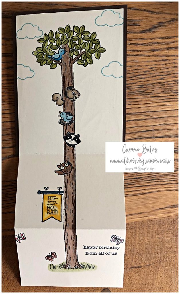 Extended out view of  children's fun fold card.  This view shows a tall tree with green leaves and a long brown trunk.  There is an owl, racoon, blue bird and squirrel face peaking out  in 4 places up the trunk.  Underneath the owl there is a yellow banner hung from the tree showing 'hip hip hooray'. At the top there is a bluebird singing on a branch.  There are blue clouds and some butterflies fluttering.  The sentiment at the bottom reads ' happy birthday from all of us'
