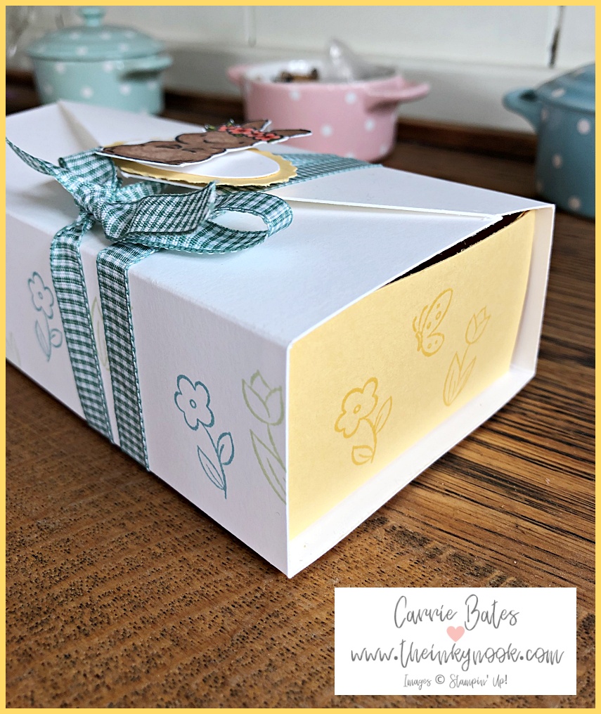 Side profile of a white handmade Easter gift box with a yellow centre wrapped in mint gingham ribbon topped with a Easter bunny wearing a floral crown