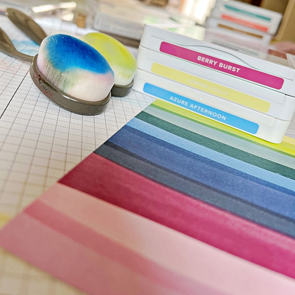 Stack of three colour ink pads next to two blending brushes and a stripy multicoloured sheet of designer series paper to make a beach hut card 