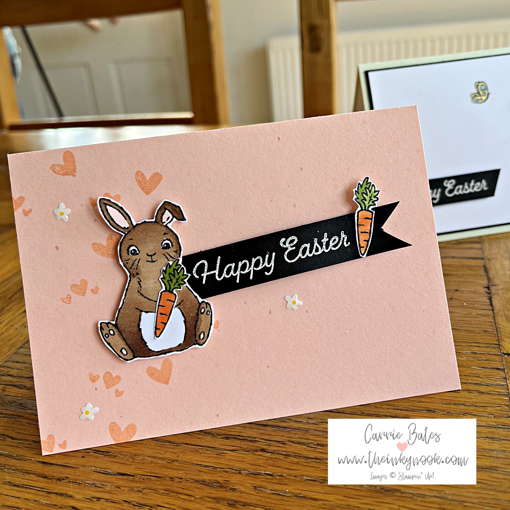 Simple Easter card on a soft pink card base stamped with soft pink hearts. A brown bunny is sitting and holding a carrot with a greeting banner reading Happy Easter in white writing on a black banner